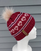 Load image into Gallery viewer, Red beanie with pink, white, and blue colorwork around in lines and heart shapes. Topped with a faux fur pom.