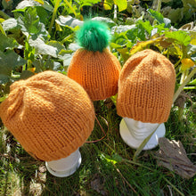 Load image into Gallery viewer, Trio of orange classic beanies sitting among pumpkin plants. Smallest beanie has a green pom.