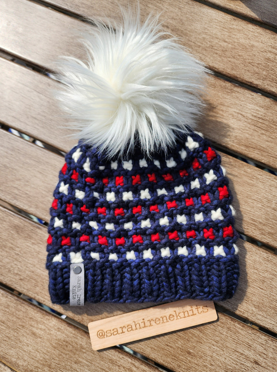 Windowpane effect beanie in navy blue with white and red rows of contrast. Topped with luxury large faux fur white pom.