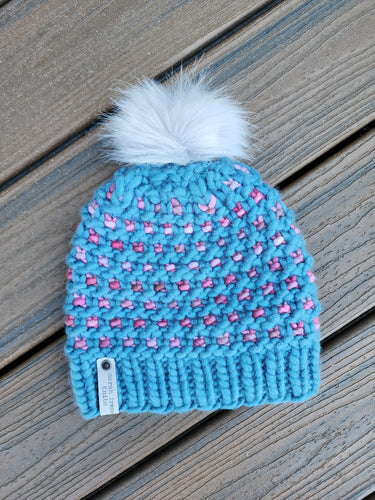 Windowpane effect beanie in teal and shades of pink. Faux fur pom on top.