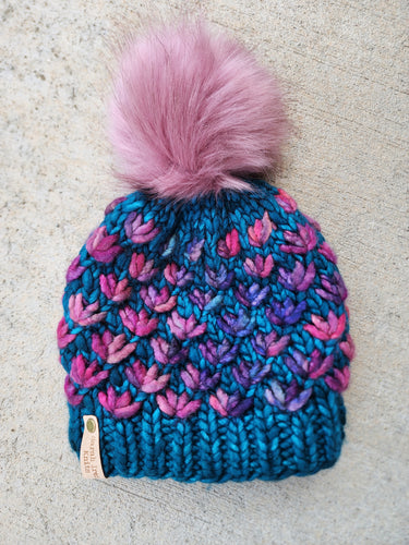 Teal beanie with purple and pink multicolor lotus flower pattern. Topped with large pink faux fur pom.