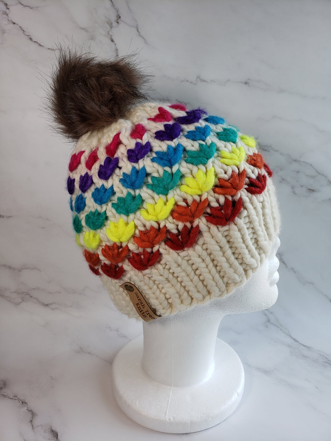 Lotus flower beanie in natural ivory wool with various rainbow colored flower rows. Brownish grey pom on top.