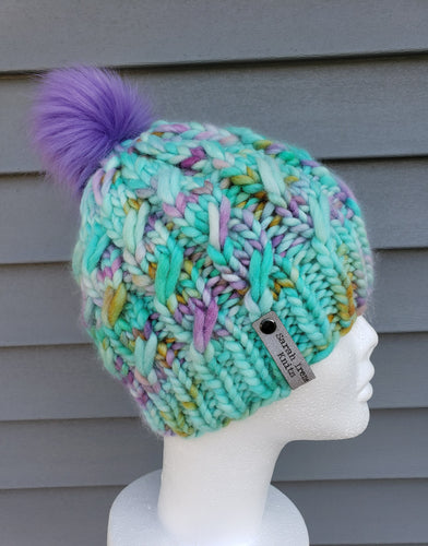 Cable effect beanie in teal with speckles of purple, yellow, and pink. Purple pom on top.