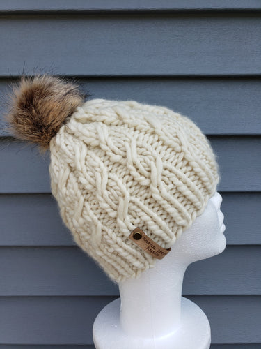 Ivory cable effect beanie with tan faux fur pom on top.