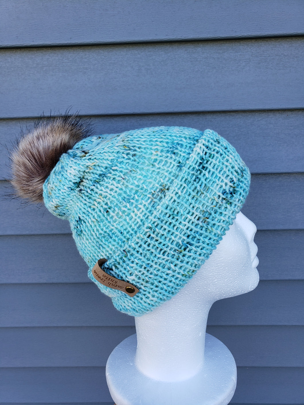 Double brim beanie in a light blue speckled yarn. Faux fur pom on top.