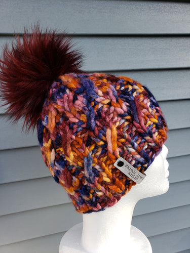 Cable effect beanie in orange, mauve, and blue multicolor. Topped with a large lux faux fur pom in burgundy.