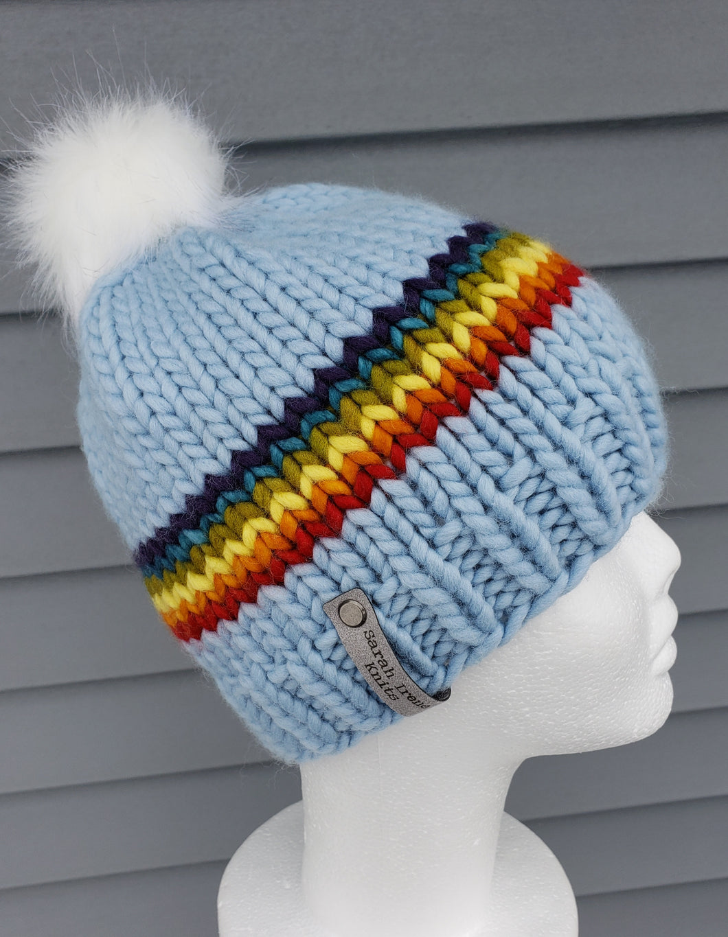 Classic beanie features a lovely rainbow stripe on sky blue background with a cloud-white faux fur pom on top.