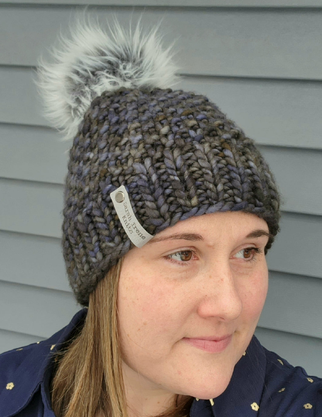 Woman wearing textured multicolor grey blue green beanie with grey faux fur pom on top.