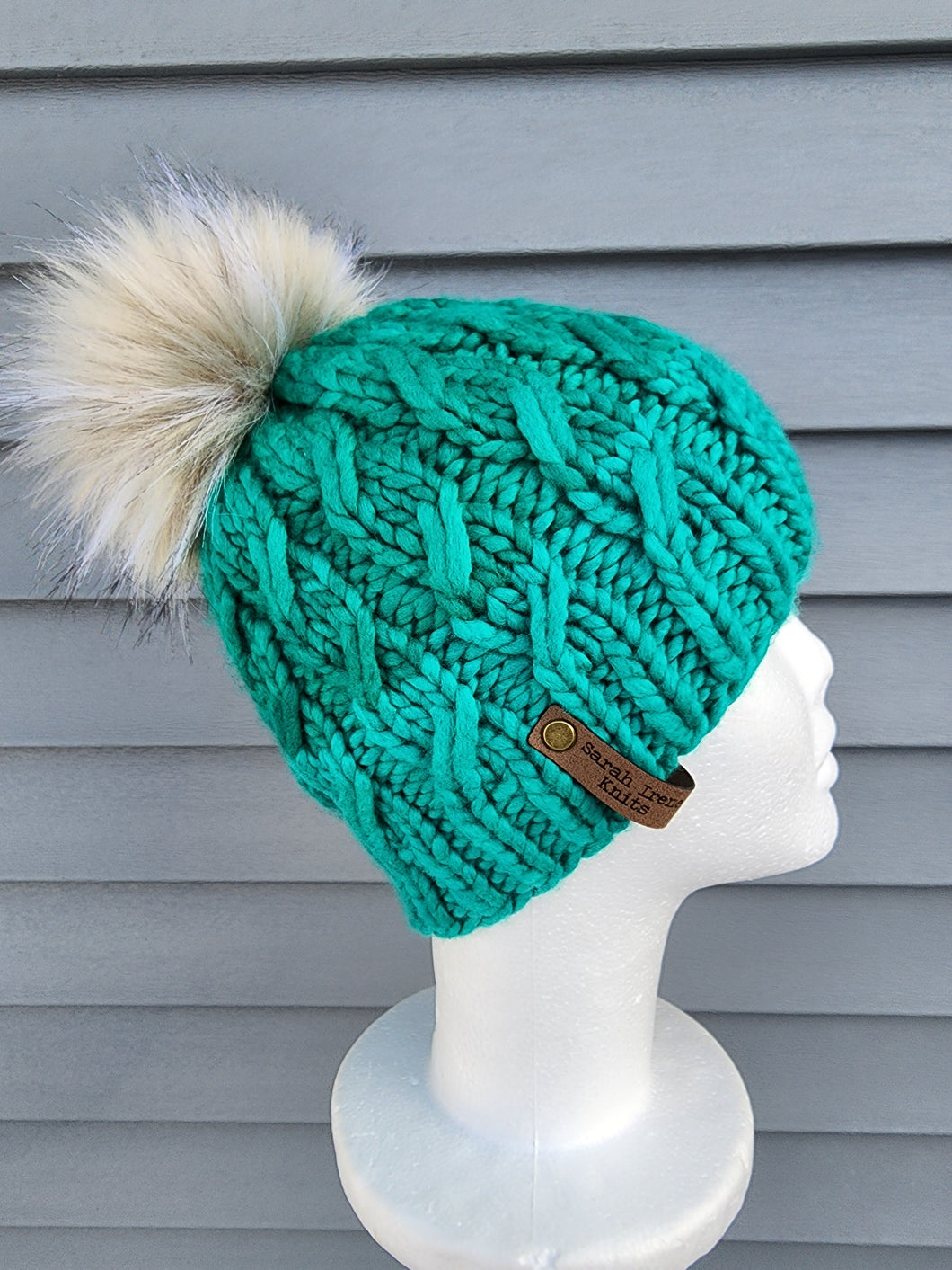 Braided effect beanie in subtle two-tone teal green color, topped with a large faux fur pom in ivory color.