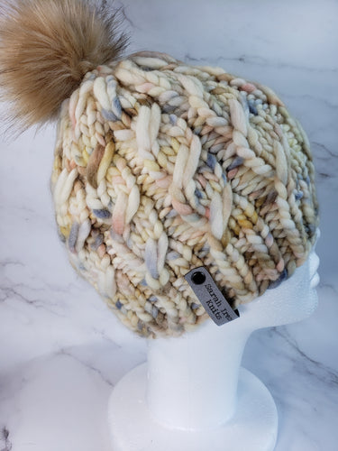 Beanie with cabling effect in a multicolor yarn with ivory, brown, blue and yellow colors topped with a faux fur pom pom