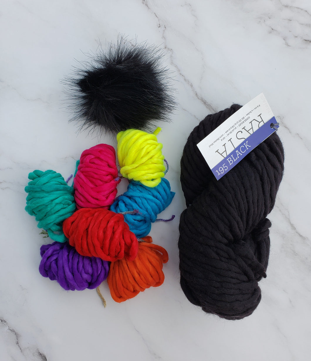 Small colorful yarn balls next to full skein of Malabrigo Rasta plus a faux fur pom.